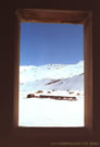 An image of a wooden window frame through which an iron bridge and snow-covered mountains can be seen, in the vicinity of Baos Morales and Baos de la Colina.